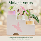 Side view of a white three-tier plant stand featuring a large pink leaf logo and a bevelled opening along its bottom edge. Pastel pots hold variegated Alocasia plants. Text invites customers to request their own logo for the side panel.