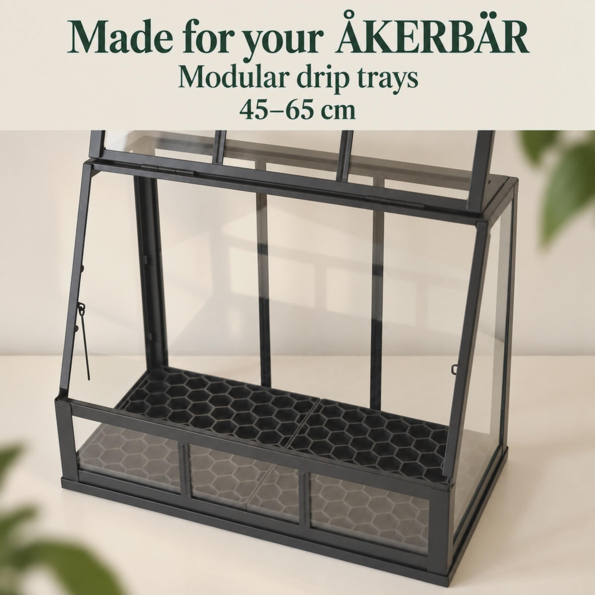 Black honeycomb drip trays fitted side by side inside an open IKEA ÅKERBÄR greenhouse. Clear panels reveal the trays beneath the removable grids, with softly blurred leaves framing the scene. Text introduces modular drip trays for 45–65 cm models.