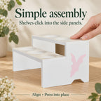 A hand presses the upper shelf of a white two-tier plant stand into place. Three circular openings sit on the lower shelf and two on the upper shelf. Solid side panels support the open-front structure, with a pink leaf logo decorating one side.